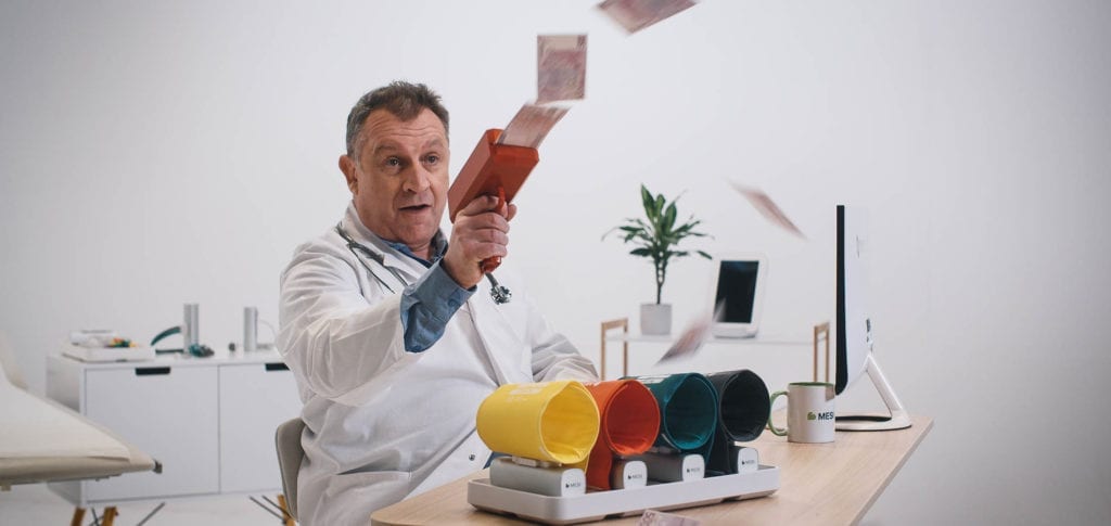 Squareme Mesi medical devices video - actor Scott Alexander Young in doctor's uniform sitting behind a desk in an office shooting notes out of a money gun on white background